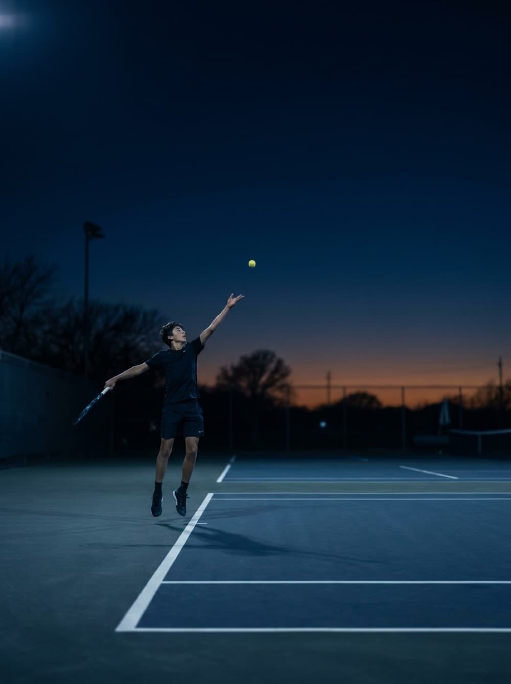 Junior player serving on a hard court at dusk